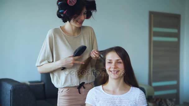 Två glada kvinnor som vänner gör roliga curlingspelare frisyr varandra och har roligt hemma — Stockvideo