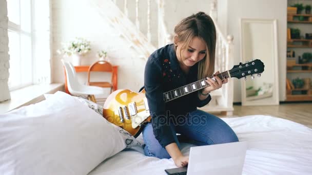 Ελκυστική νεαρή κοπέλα που μαθαίνουν να παίζουν κιθάρα ηλεκτρική με σημειωματάριο κάθονται στο κρεβάτι στο υπνοδωμάτιο στο σπίτι — Αρχείο Βίντεο