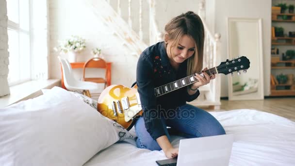 Jovem atraente aprendendo a tocar guitarra elétrica com notebook sentar na cama no quarto em casa — Vídeo de Stock