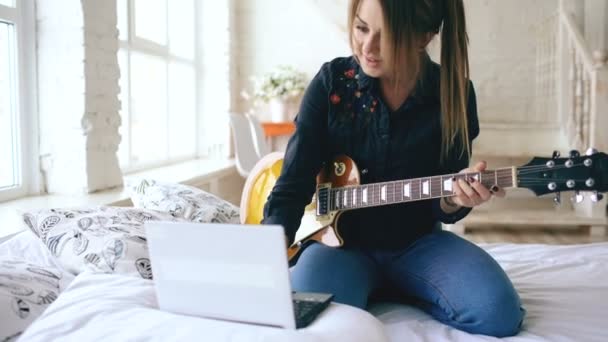 Jovem atraente aprendendo a tocar guitarra elétrica com notebook sentar na cama no quarto em casa — Vídeo de Stock