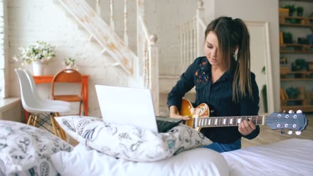 Привлекательная молодая девушка учится играть на электрогитаре с ноутбуком сидеть на кровати в спальне дома — стоковое видео