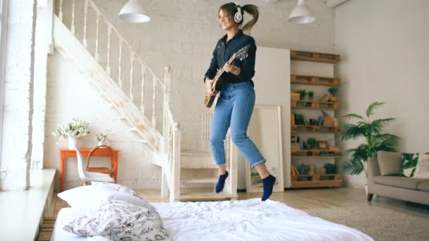 Grappige jonge vrouw hoofdtelefoon springen op het bed met elektrische gitaar in slaapkamer thuis binnenshuis — Stockvideo