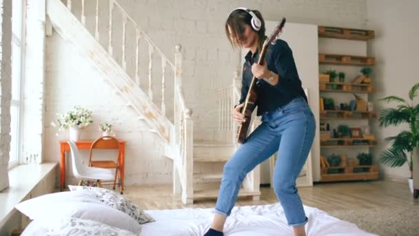 Jeune femme drôle dans les écouteurs jouer de la guitare électrique sur le lit dans la chambre à coucher à la maison à l'intérieur — Video