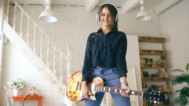Vacker ung leende flicka i hörlurar poserar med elgitarr hemma studio inomhus — Stockvideo