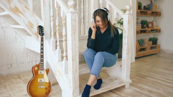 Gyönyörű fiatal nő, a lépcsőn ülve hallgatni zenét otthon fejhallgató beltéri — Stock videók