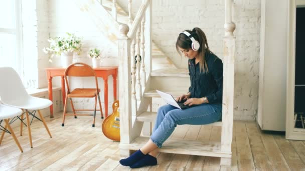 Hermosa mujer con auriculares que se relaja con el ordenador portátil mientras está sentado en las escaleras en la sala de estar en casa — Vídeo de stock