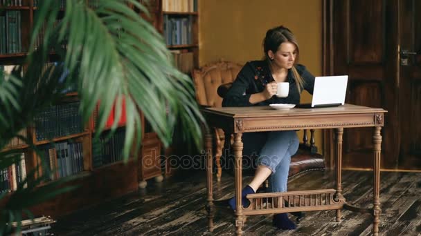 Lächelndes Studentenmädchen arbeitet am Laptop und trinkt in der Universitätsbibliothek drinnen Kaffee — Stockvideo