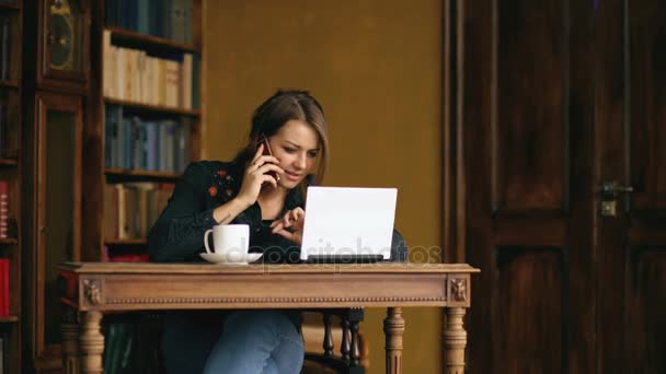 Chica estudiante bastante joven hablando en el teléfono móvil mientras estudia en la biblioteca en el interior — Vídeo de stock