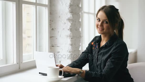 Mosolygó fiatal diák nő ül asztalhoz a laptop beltéri kávézóban — Stock videók