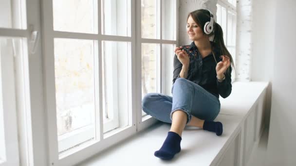 Mujer joven con auriculares escuchando música y bailando sentarse en la ventana en el interior — Vídeos de Stock