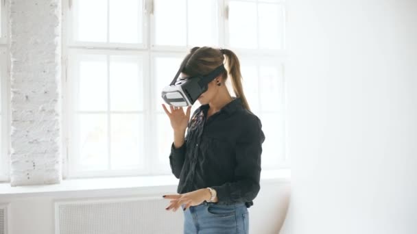 Mulher no fone de ouvido VR olhando para cima e tentando tocar objetos na realidade virtual na sala branca dentro de casa — Vídeo de Stock