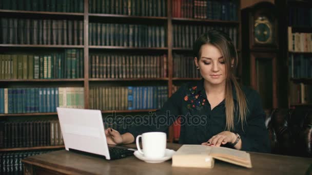 Mosolygó diák lány dolgozik laptop és olvas könyvet, az egyetemi könyvtár beltéri — Stock videók