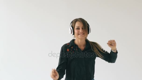 Cámara lenta de mujer joven en auriculares escuchando música y bailando sobre fondo blanco en interiores — Vídeo de stock
