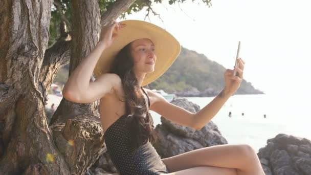 Молодая сексуальная женщина в купальнике и шляпе делает селфи съемки с помощью смартфона во время отдыха на пляже — стоковое видео
