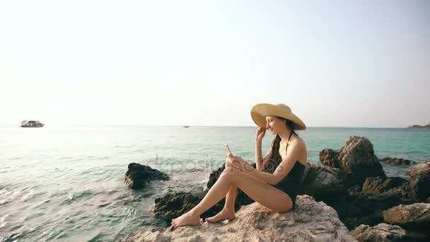 Молодая сексуальная женщина в купальнике и шляпе делает селфи съемки с помощью смартфона во время отдыха на пляже — стоковое видео