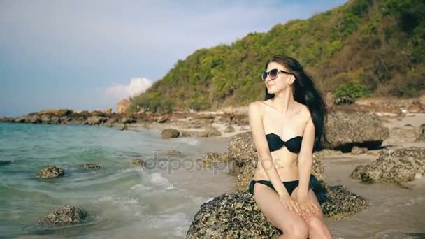 Young sexy woman in sunglasses smiling and sitting on rock at beautiful tropical beach — Stock Video