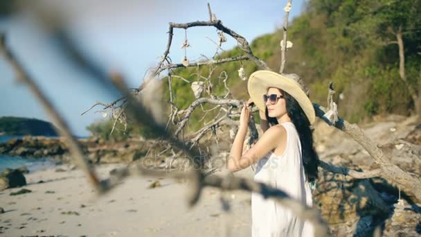 Jovem mulher sexy em óculos de sol e chapéu sorrindo e posando perto da árvore na bela praia tropical — Vídeo de Stock