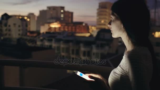 Close-up de jovem mulher sorridente mensagens de texto no smartphone em pé no terraço do último piso à noite — Vídeo de Stock