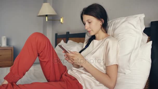 スマート フォンと笑顔をしてホテルの部屋でベッドに横になっている金髪女性をかなり — ストック動画