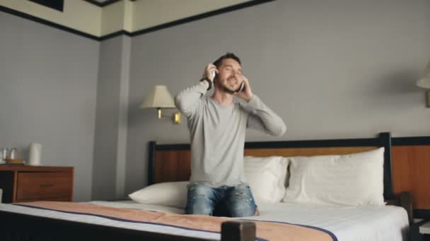 Jonge gelukkig man luisteren naar muziek met draadloze hoofdtelefoons en dansen op bed in hotelkamer — Stockvideo