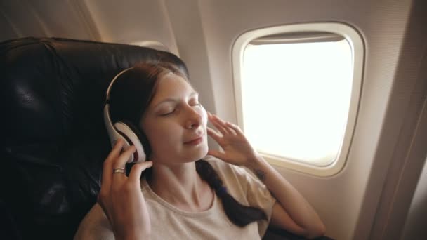 Młoda kobieta w słuchawki bezprzewodowe słuchanie muzyki i uśmiechając się w locie samolotem — Wideo stockowe