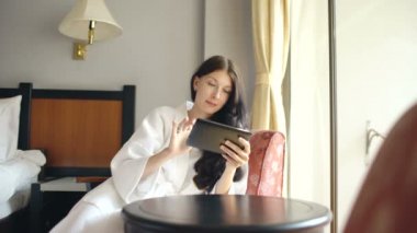 Otel odasında sandalye üzerinde oturan tablet bilgisayar kullanarak bornoz içinde genç iş kadını