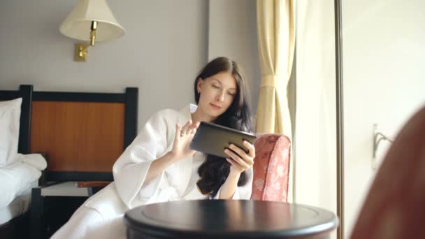 Jeune femme d'affaires en peignoir à l'aide d'une tablette assise sur une chaise dans la chambre d'hôtel — Video