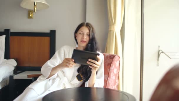 Гарненька молода жінка в халаті спілкується на планшетному комп'ютері, сидячи на стільці в готельному номері — стокове відео