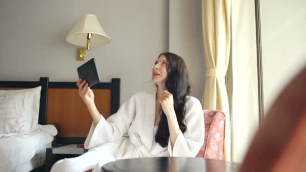 Csinos, fiatal nő tabletta számítógép ül a székre, a szállodai szobában fürdőköpeny beszélget — Stock videók