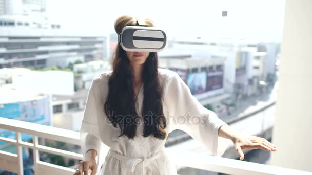 Mulher de roupão de banho vestindo fone de ouvido realidade virtual em pé na varanda do quarto do hotel e tem experiência VR — Vídeo de Stock