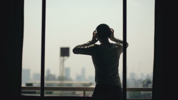 Silhouet van jonge man ad luisteren muziek dansen in wireles hoofdtelefoon stand op hotel kamer balkon — Stockvideo