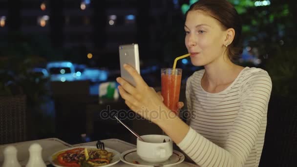 Gelukkige vrouw schieten selfie met smartphone en drinck cocktail juice zittend in restaurant nachts — Stockvideo