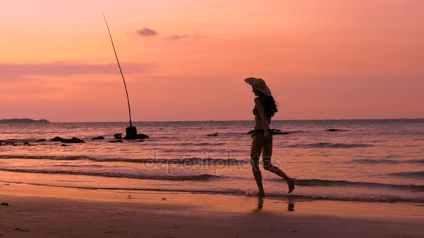 Медленно движущийся силуэт счастливой женщины в шляпе, бегущей по пляжу во время заката на отдыхе — стоковое видео