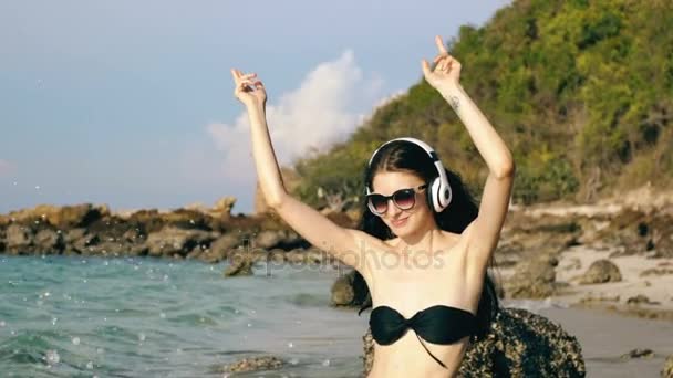 Cámara lenta de la hermosa mujer feliz escuchando música en los auriculares inalámbricos y bailando en la playa del océano — Vídeo de stock
