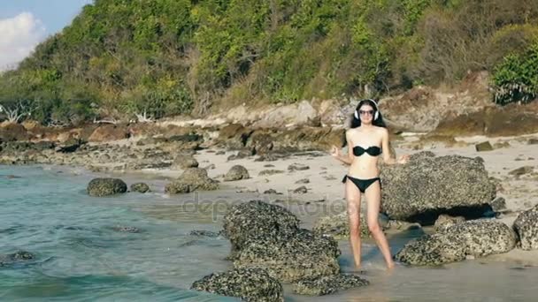 Cámara lenta de la hermosa mujer feliz escuchando música en los auriculares inalámbricos y bailando en la playa del océano — Vídeos de Stock