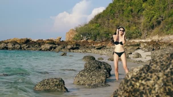 Cámara lenta de la hermosa mujer feliz escuchando música en los auriculares inalámbricos y bailando en la playa del océano — Vídeos de Stock