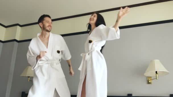 Slow motion van jonge gelukkige paar in badjas springen en dansen op bed in het hotel tijdens hun huwelijksreis vakantie — Stockvideo