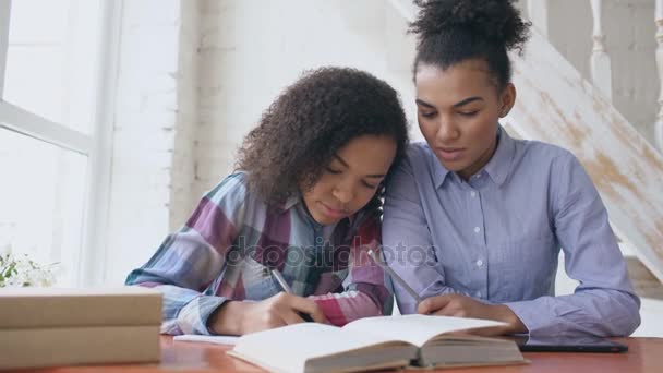 Junges Mädchen mit lockigem Haar und gemischter Rasse sitzt am Tisch und konzentriert sich auf den Unterricht und ihre ältere Schwester hilft ihr beim Lernen — Stockvideo