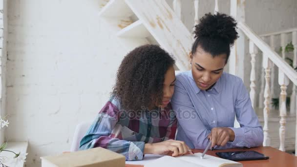 Adolescente de pelo rizado mestizo chica joven sentada en la mesa concentrándose lecciones de aprendizaje enfocadas y su hermana mayor la ayuda a estudiar — Vídeos de Stock