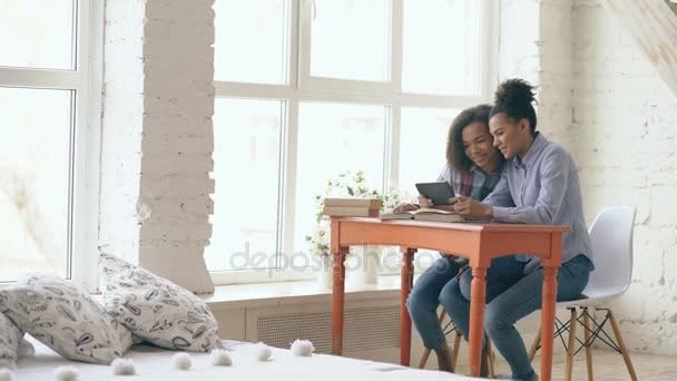 Две симпатичные кудрявые девушки смешанной расы, сидящие за столом, веселятся, изучая уроки и используя тарелку дома — стоковое видео