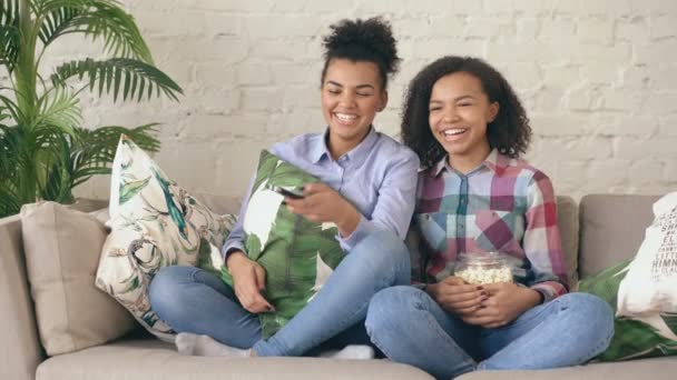 Deux copines frisées de race mixte assises sur le canapé et regardent des films comiques et mangent du pop-corn. Sœurs drôles se détendre ensemble à la maison — Video