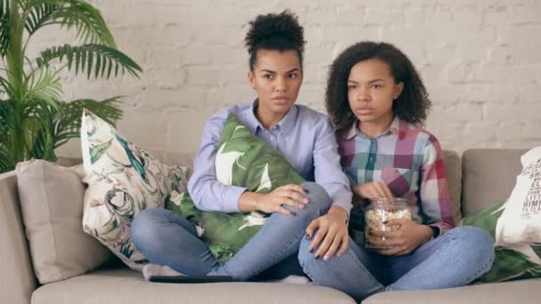 Två blandad ras curly girl vänner sitter på soffan och titta på thriller film och äter popcorn hemma — Stockvideo