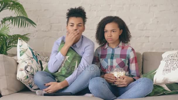 Δύο μικτή φυλή σγουρά κορίτσι φίλοι κάθονται στον καναπέ και να παρακολουθήσουν νευρικό τηλεόραση δείχνουν και να τρώνε ποπ κορν στο σπίτι — Αρχείο Βίντεο