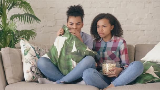 Two mixed race curly girl friends sitting on the couch and watch very scary movie on TV and eat popcorn at home — Stock Video