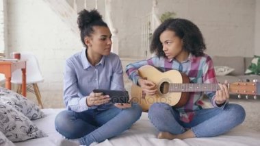 Akustik gitar evde oynamak için genç kız kardeşi öğretim yatağa oturmuş tablet bilgisayar ile karışık yarış genç kadın