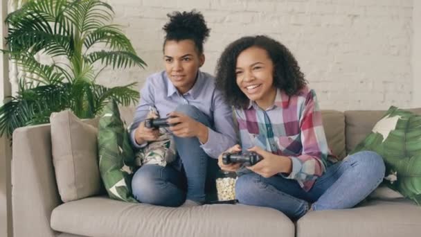 Dos amigos de la chica rizada de raza mixta sentados en el sofá juegan juegos de computadora de consola con mando y se divierten en casa — Vídeo de stock