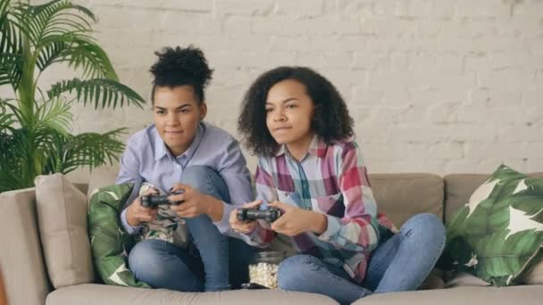 Twee gemengd ras krullend vriendinnen zittend op Bank console computerspelletjes spelen met gamepad en veel plezier thuis — Stockvideo