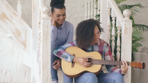 Deux filles frisées brésiliennes sœurs assises sur les escaliers et s'entraînent à jouer de la guitare acoustique. Les amis s'amusent et chantent à la maison — Video