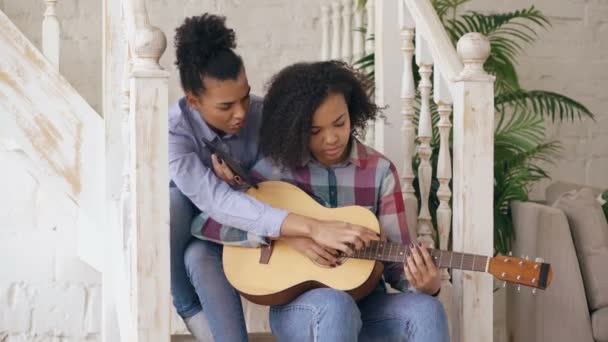 Mixte jeune femme assise dans les escaliers enseignant à sa sœur adolescente à jouer de la guitare acoustique à la maison — Video