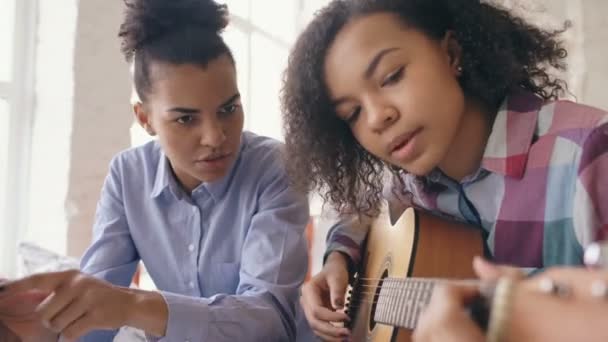Jovem de raça mista com computador tablet sentado na cama ensinando sua irmã adolescente a tocar guitarra acústica em casa — Vídeo de Stock